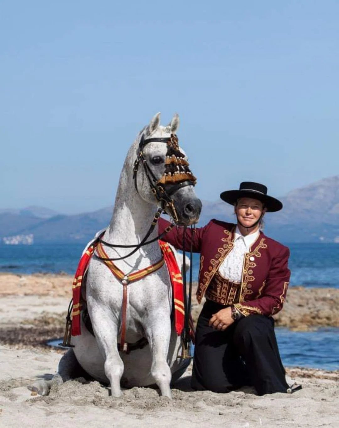 A grey horse in traditional Mallorcan tack with a rider in period costume by the sea