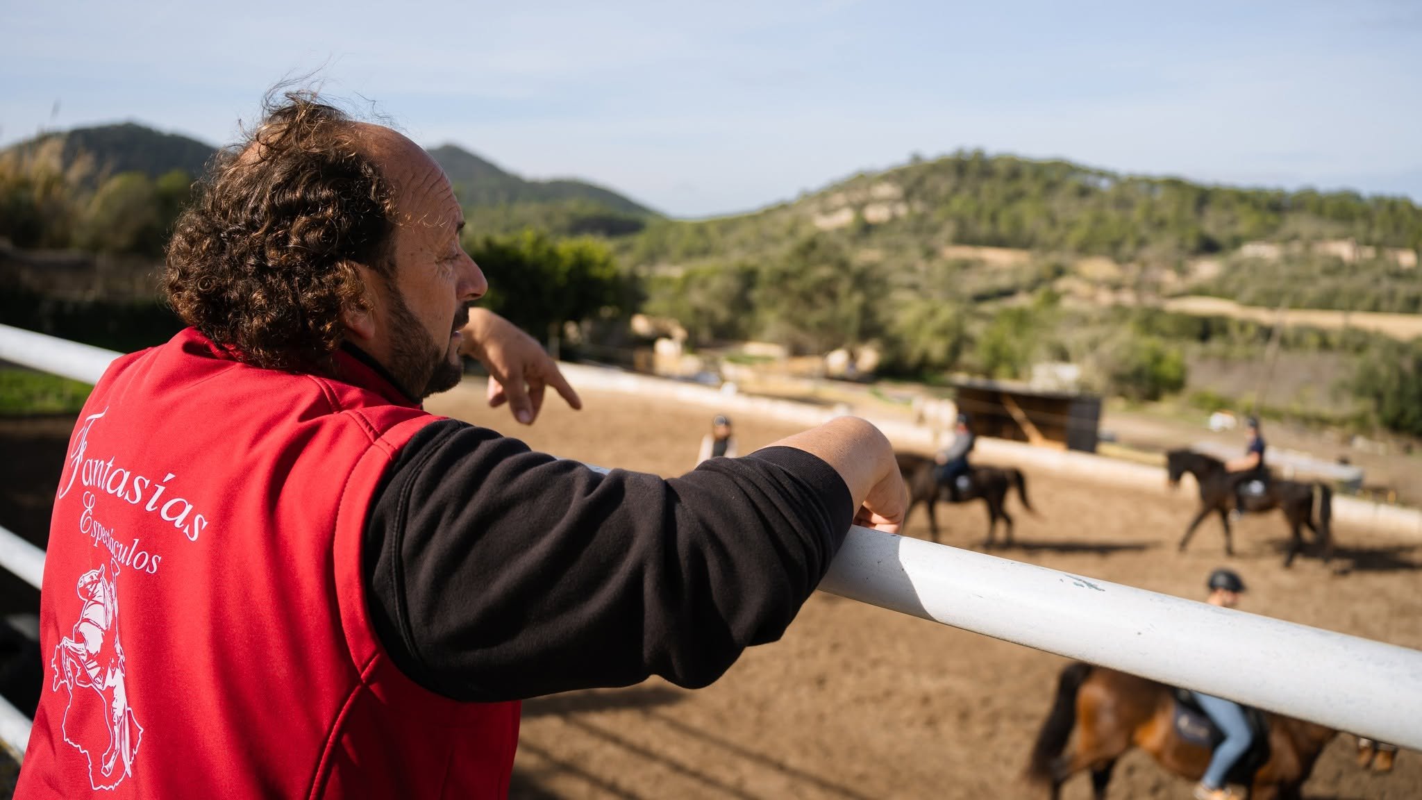 An instructor leans on the arena rail and watches a lesson