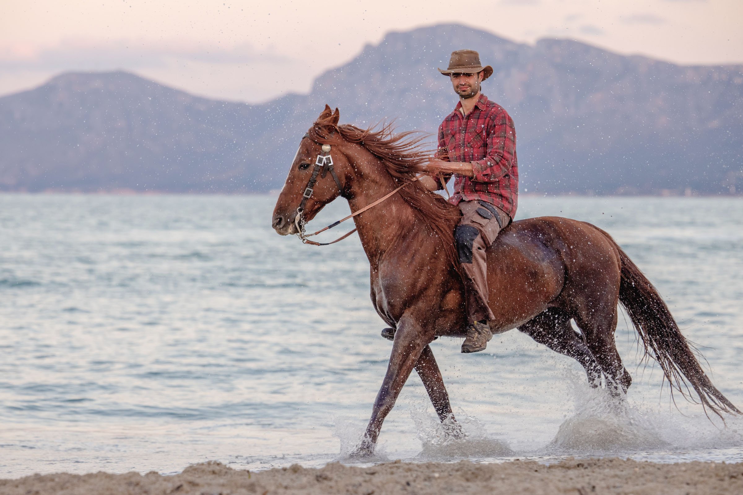 A rider cantering a chestnut horse through shallow sea, mountains behind