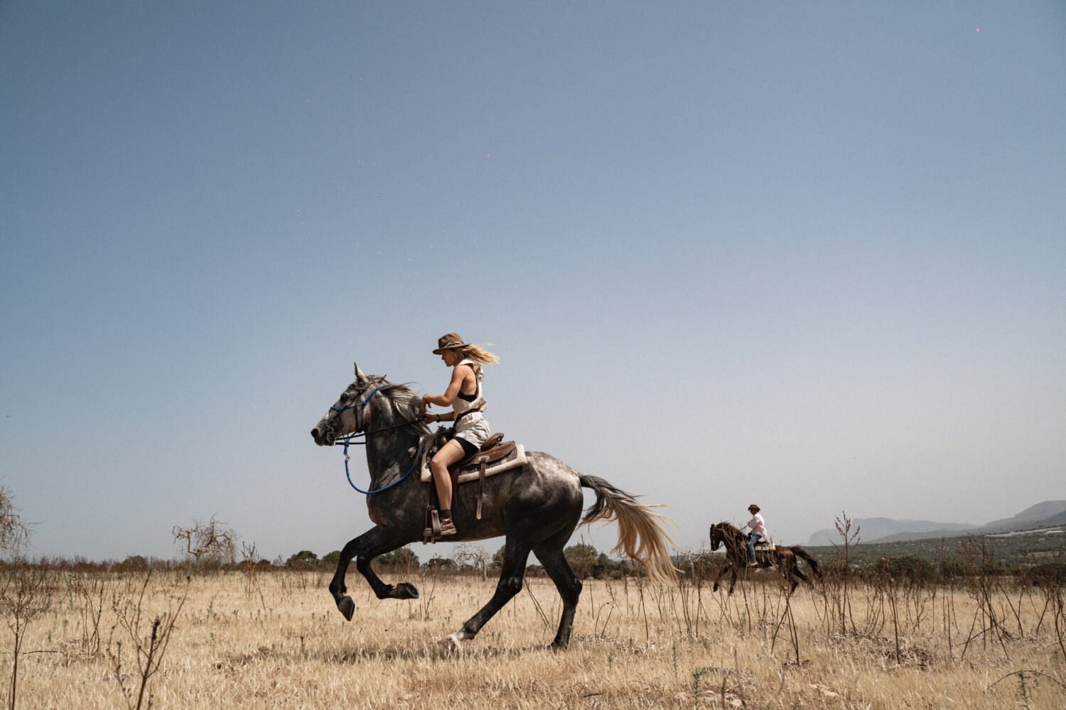 A rider galloping a grey horse across a dry field, a second rider behind
