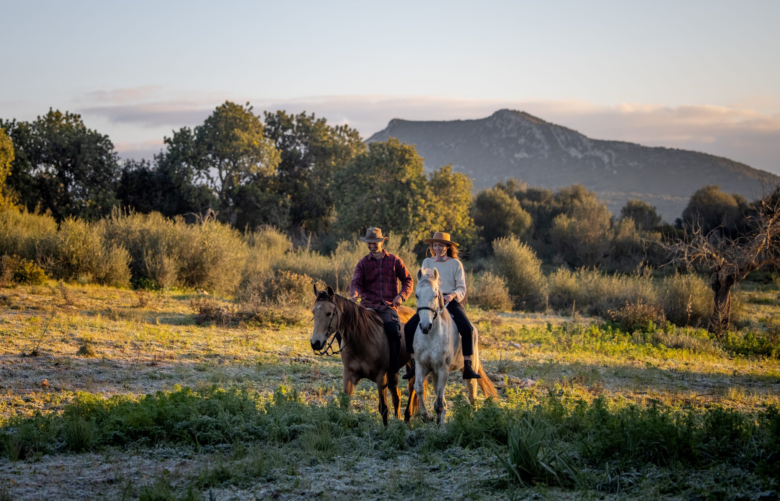 Two riders on a bay and a grey horse in evening light, a hill behind